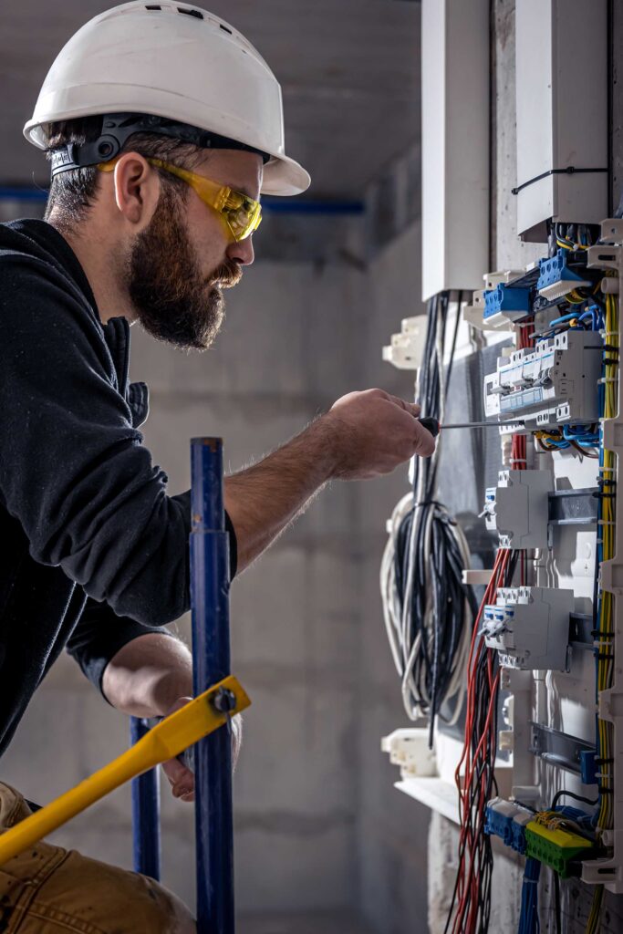 A construction worker in a white hard hat and safety glasses uses a screwdriver on an electrical panel. He appears focused in a partially constructed room.