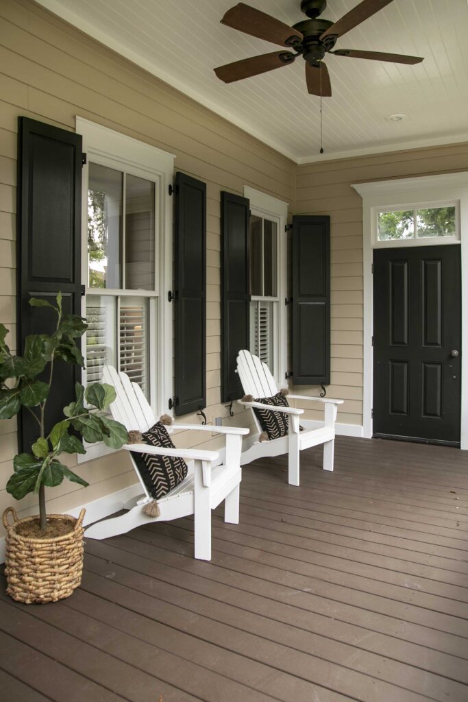A cozy porch featuring two white Adirondack chairs with black geometric pillows, dark wood floor, beige walls, black shutters, a ceiling fan, and a potted plant.
