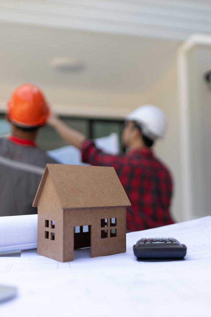 A small house model sits on blueprints with a calculator and tape measure. Two workers in helmets discuss construction in the blurred background.